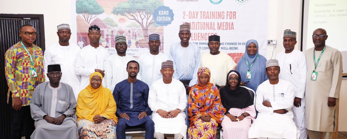 A group of 16 people, dressed in traditional and semi-formal attire, pose for a photo in front of a white banner. The banner has the text "2-DAY TRAINING FOR TRADITIONAL MEDIA INFLUENCERS" and logos for EngenderHealth and other organizations. The people are arranged in two rows, with some standing and others seated. Most of the individuals are smiling and looking towards the camera. The background is a plain wall with some light fixtures visible.