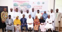 A group of 16 people, dressed in traditional and semi-formal attire, pose for a photo in front of a white banner. The banner has the text "2-DAY TRAINING FOR TRADITIONAL MEDIA INFLUENCERS" and logos for EngenderHealth and other organizations. The people are arranged in two rows, with some standing and others seated. Most of the individuals are smiling and looking towards the camera. The background is a plain wall with some light fixtures visible.